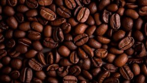 Café nas refeições e seus efeitos na saúde "Close-up de grãos de café torrado, com textura rica e cor marrom intensa. A imagem representa o consumo de café durante ou após as refeições – um hábito comum que pode impactar a absorção de nutrientes, a digestão e a saúde gastrointestinal. Ideal para conteúdos sobre nutrição, saúde digestiva e hábitos alimentares."